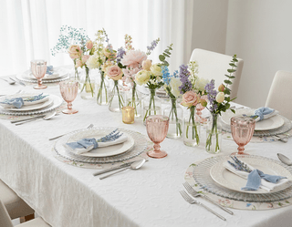 Table setting with floral arrangements, floral plates, blue napkins, pink glasses, and white textured tablecloth.