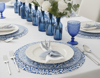 Table setting with blue patterned placemats, white plates, blue glasses and bottles with white roses.