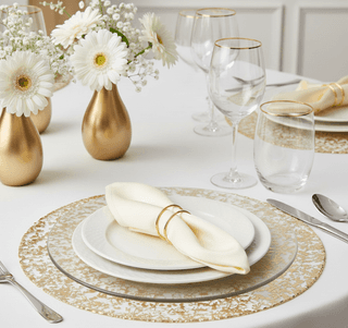 Elegant table setting: white plates, gold-rimmed glasses, floral vases, & gold-flecked placemats on a white tablecloth.