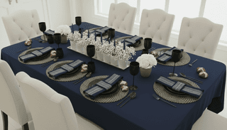 Formal dining table setting with navy tablecloth, black glassware, patterned chargers, and cream-colored chairs.