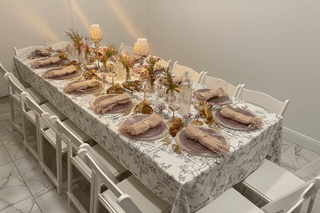 Elegant dining table set with floral tablecloth, pink plates and napkins, candles, and white chairs.