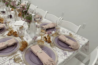 A table set for a meal with purple plates, pink napkins, gold silverware, & flower arrangements.