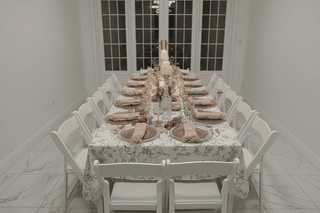 Formal dining table with floral tablecloth, pink place settings, candles, and white chairs.