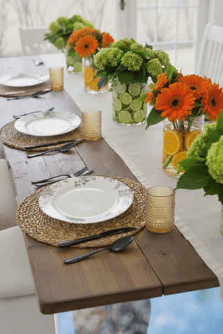 Wood table set with plates on woven mats, black flatware, patterned tumblers, and vases with citrus slices and flower arra...